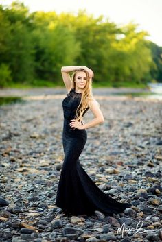 a woman in a long black dress standing on rocks with her hands behind her head