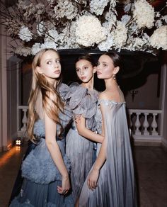 three young women standing next to each other in front of a floral arrangement with white flowers