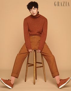 a man sitting on top of a wooden stool wearing brown pants and a turtle neck sweater