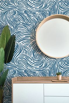 a mirror on the wall next to a dresser and plant in a room with blue patterned walls