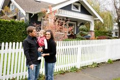 a man and woman standing in front of a white picket fence with a baby on her lap