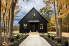 a small black house surrounded by trees and grass with fall foliage on the ground around it