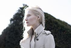 a woman with blonde hair wearing a white coat and looking off into the distance while standing in front of some bushes