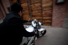 a man riding on the back of a motorcycle down a street next to a wooden wall