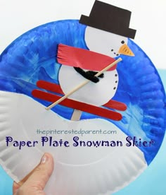 a paper plate snowman with a hat and skis on it, is held up by a hand