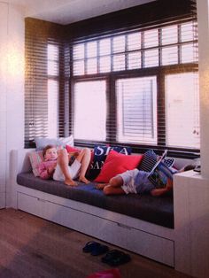 two children are laying on a couch in front of a window with shutters and pillows