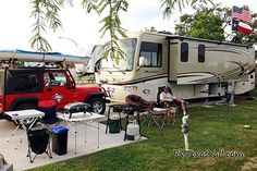 an rv parked in the grass next to a red truck and camper with two people sitting on chairs