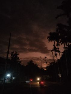 cars driving down the road at night with palm trees on both sides and dark clouds in the background