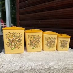 four yellow flowered containers sitting on top of a white cloth covered tablecloth next to a window