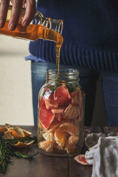 a person pouring orange juice into a jar filled with sliced up citrus fruit and herbs