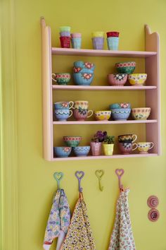 a shelf filled with cups and teacups on top of hooks next to a yellow wall