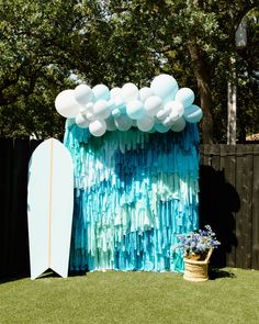 a surfboard sitting in the grass next to a wall with blue and white balloons