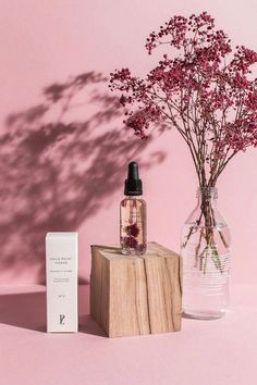 a vase with flowers in it sitting next to a box and a bottle on a pink surface