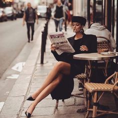 a woman is sitting at an outdoor table reading a newspaper while looking down the street