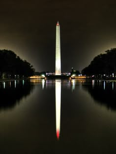 the washington monument is lit up at night with its reflection in the water and trees around it