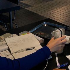 a person sitting at a table with papers and pens in front of them, holding a cup