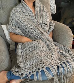 a woman sitting on a couch wearing a gray crocheted shawl with fringes