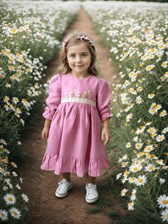 girl wearing a pink linen long-sleeved dress with hand-embroidered daisy flowers on the front poses in a field of daisies Pink Princess Style Embroidered Dress, Pink Linen Ruffled Dress, Pink Embroidered Dress For Dress-up Occasions, Cute Pink Dress With Floral Embroidery, Pink Embroidered Dress For Spring, Pink Embroidered Dress With Hem Detail For Spring, Pink Embroidered Dress With Embroidered Hem For Spring, Pink Floral Embroidery Dress For Baptism, Pink Long Sleeve Dress With Embroidered Hem