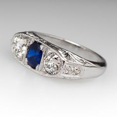 This lovely circa 1930s antique sapphire ring is centered with one (1), bead set, oval cut natural blue sapphire weighing 0.81 carats and flanked to each side by one (1), bead set, old European cut diamond. The shoulders of the ring are each bead set with two (2) round single cut diamonds. The gemstones are bordered with milgrain edging. Engraved details accent the side faces of the ring. The ring measures 7.3mm at the top, rises 3.7mm above the finger, tapering to 2.9mm wide and 1.6mm thick at the base of the shank. The ring is currently a size 6 and we offer complimentary resizing to fit. Classic Oval Sapphire Ring, Classic Sapphire Birthstone Ring, Classic Sapphire Halo Ring, Antique Oval Three Stone Diamond Ring, Blue Oval Sapphire Ring In Art Deco Style, Art Deco Blue Oval Sapphire Ring, Antique Blue Oval Diamond Ring, Art Deco Oval Sapphire Ring In Platinum, Art Deco Oval Sapphire Ring With Center Stone