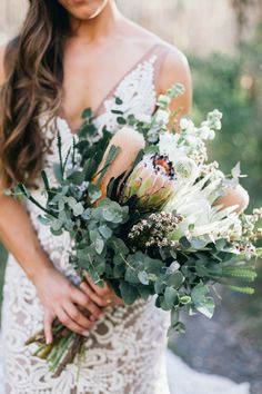 a woman holding a bouquet of flowers and greenery in her hands while wearing a wedding dress