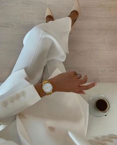 a woman wearing a white dress and gold watch with her hands on the table next to a cup of coffee