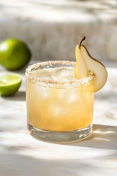 a glass filled with an alcoholic drink next to two limes and a slice of apple