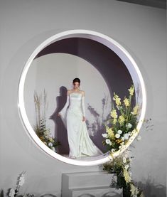 a woman in a white dress is standing behind a circular mirror with flowers on it