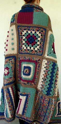 a woman wearing a multicolored crocheted granny afghan shawl standing in front of a white wall