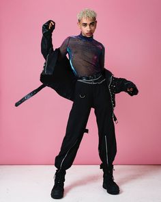 a woman with blonde hair and black pants posing in front of a pink background wearing a costume