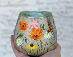 a hand holding a painted vase in front of a white wall with flowers and butterflies on it