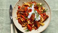 a white bowl filled with pasta and meat covered in sauce next to two silverware