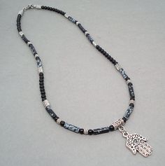 "This distinctive necklace features a metal pendant of Hamsa, known as an amulet shaped like a hand and is thought to protect against the evil eye. This detailed Hamsa charm is a made-in-USA high quality TierraCast product, which is made of pewter in antiqued fine silver finish. It hangs from a necklace that I designed with natural Snowflake Obsidian stone tube beads in black with whitish gray patches resembling snowflakes, and Black Onyx gemstone round beads. I have also added antiqued silver f Black Amulet Necklaces With Polished Beads, Black Amulet Necklace With Polished Beads, Black Amulet Style Necklaces With Polished Beads, Black Amulet Beaded Necklaces For Jewelry Making, Black Amulet Style Beaded Necklaces, Hand Of Hamsa, Hamsa Charm, Mens Necklace, Obsidian Stone