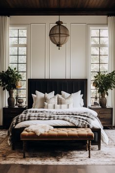 a bedroom with white walls and wood flooring has a black headboard, plaid blanket, and footstool