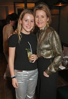 two women standing next to each other at an event, one holding a drink and the other smiling