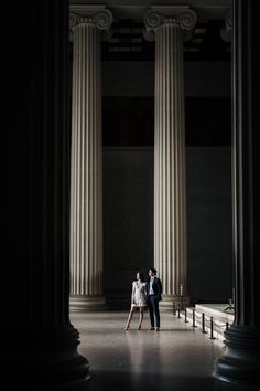 an engaged couple standing in front of two large pillars at the end of a hallway