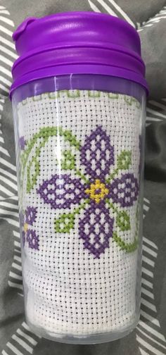 a purple and white cross stitched cup with a purple lid sitting on top of a bed