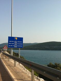 a blue sign on the side of a road next to a body of water with hills in the background