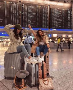 two women sitting on suitcases at an airport posing with their arms in the air