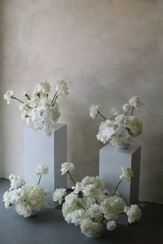 three white vases with flowers in them sitting on a gray surface next to a window