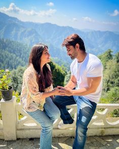 a man kneeling down next to a woman on top of a stone wall with mountains in the background