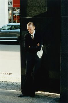 a man in a suit leaning against a black wall on the side of a street