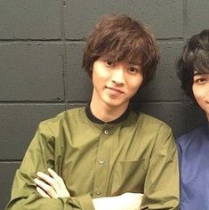 two young men standing next to each other in front of a gray wall with their arms crossed