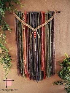 a wall hanging made out of different colored yarns and beads with plants in the background