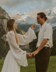 a man and woman standing on top of a lush green field next to a mountain