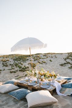 a table set up on the beach with pillows and an umbrella