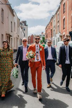 Groom in orange suit with patterned tie and white flower buttonhole and sunglasses walking with groomsmen in suits and sunglasses for Bristol city wedding Mens Colored Suits Wedding, Non Traditional Suit Men, Men’s Wedding Suits 2023, Wedding Suits Men Colourful, Funky Wedding Suits Men, Bright Groom Suit, Mens Mismatched Suits, Coloured Wedding Suit, Coloured Groom Suit