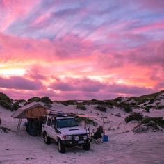 a white truck parked on top of a sandy beach under a pink and purple sky