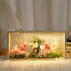 an arrangement of flowers in a glass case on a white furnishing next to a lamp