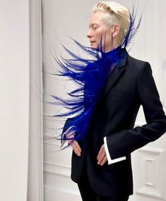 a woman in a black suit with blue feathers on her head and hair blowing in the wind