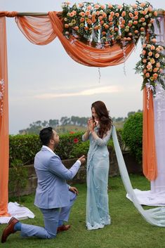 a man kneeling down next to a woman in front of an orange and white wedding arch